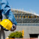 A man in blue jeans and a hard hat holding a yellow helmet, ready for security needs assessment to improve building safety